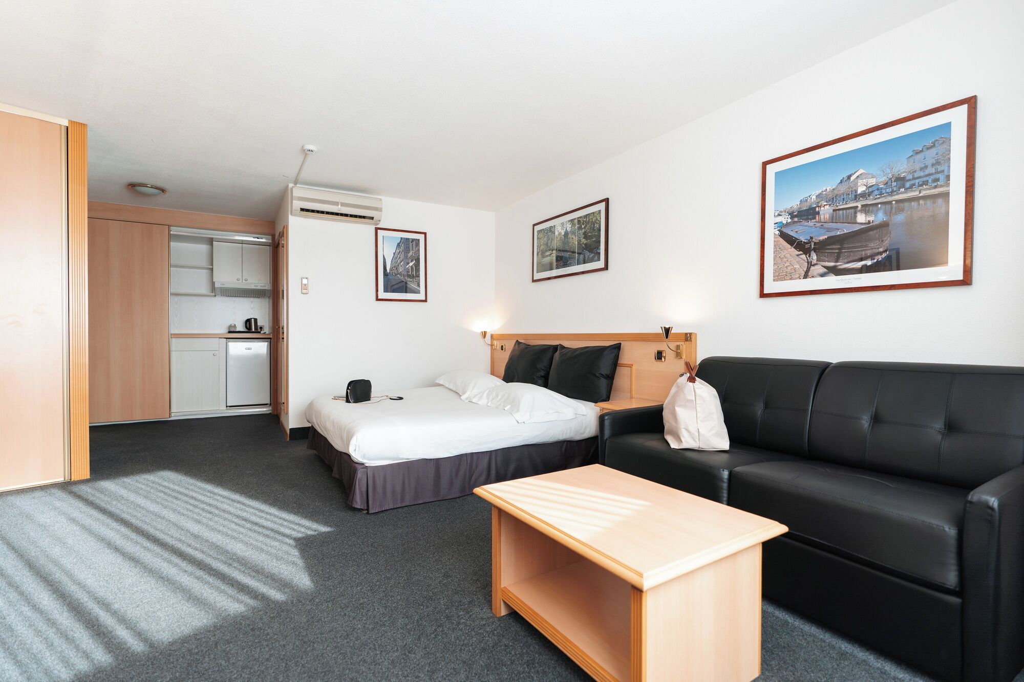 A hotel room with a bed, a sofa, a small wooden table, a kitchenette, and a bathroom.