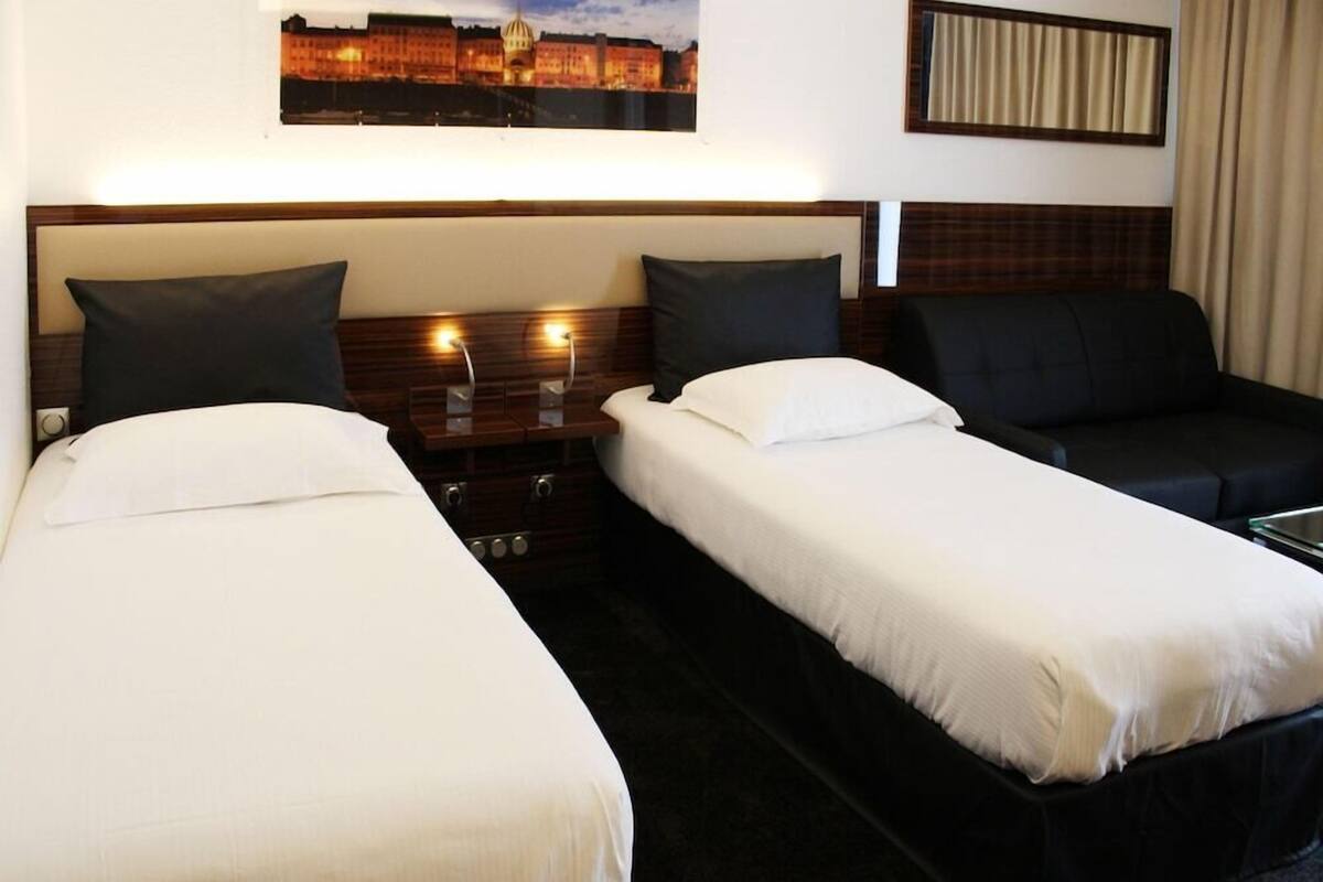 A hotel room with two single beds, a nightstand with a lamp, a sofa, and a cityscape painting on the wall.