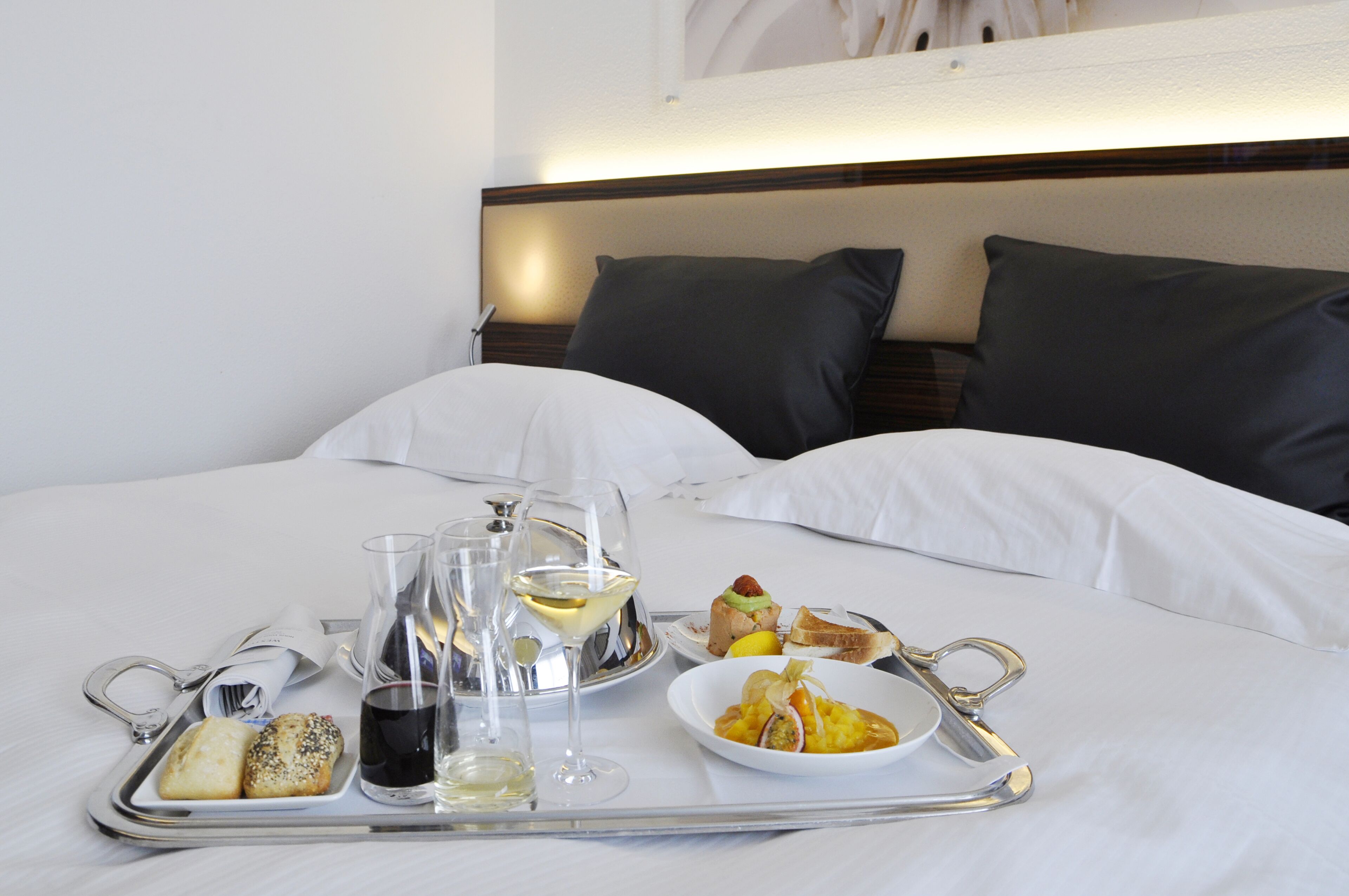 Una cama bien tendida con sábanas blancas, una bandeja con comida y bebidas, y un cabecero con almohadas oscuras.