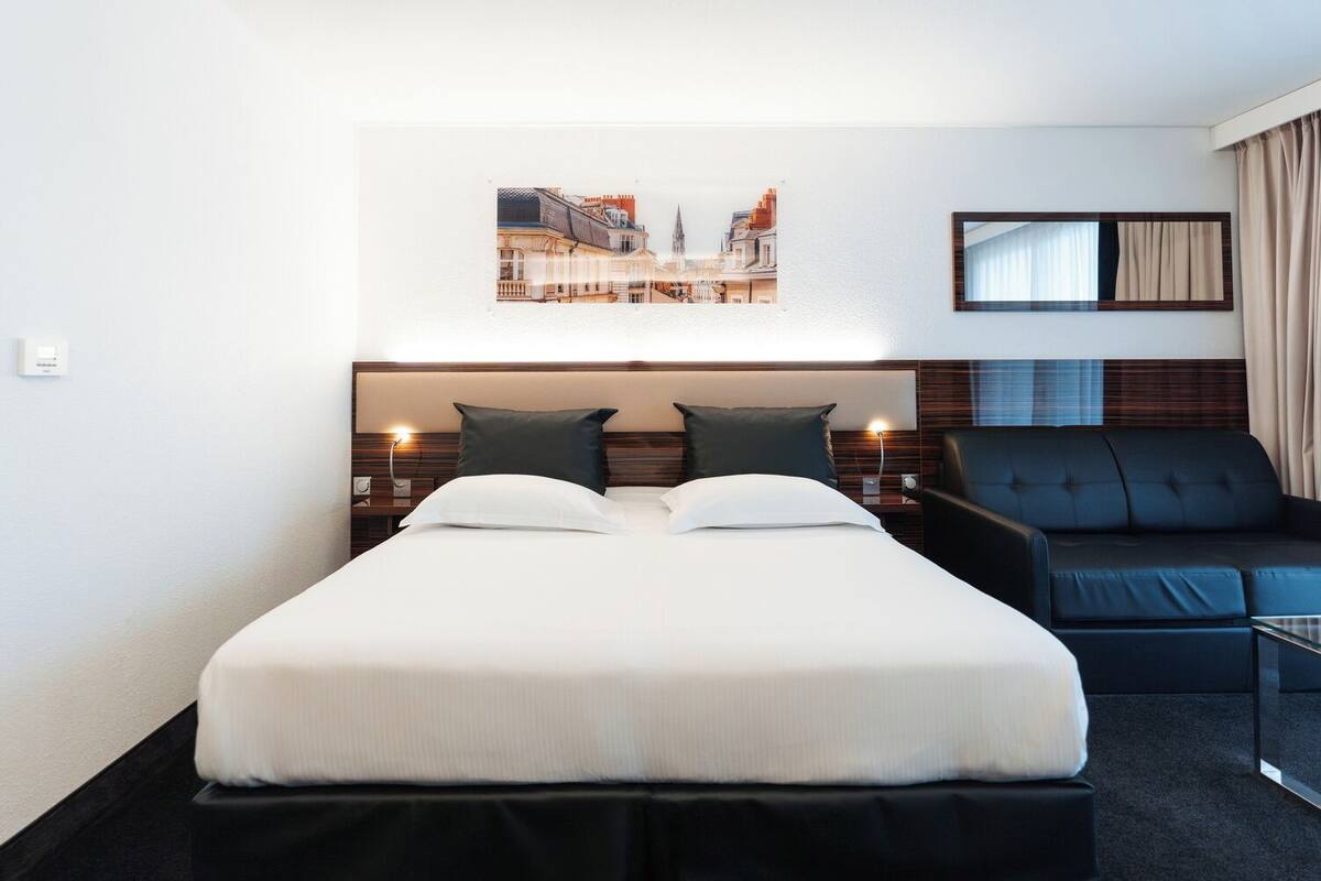 A hotel room with a double bed, a sofa, a wall-mounted TV, and a cityscape artwork.