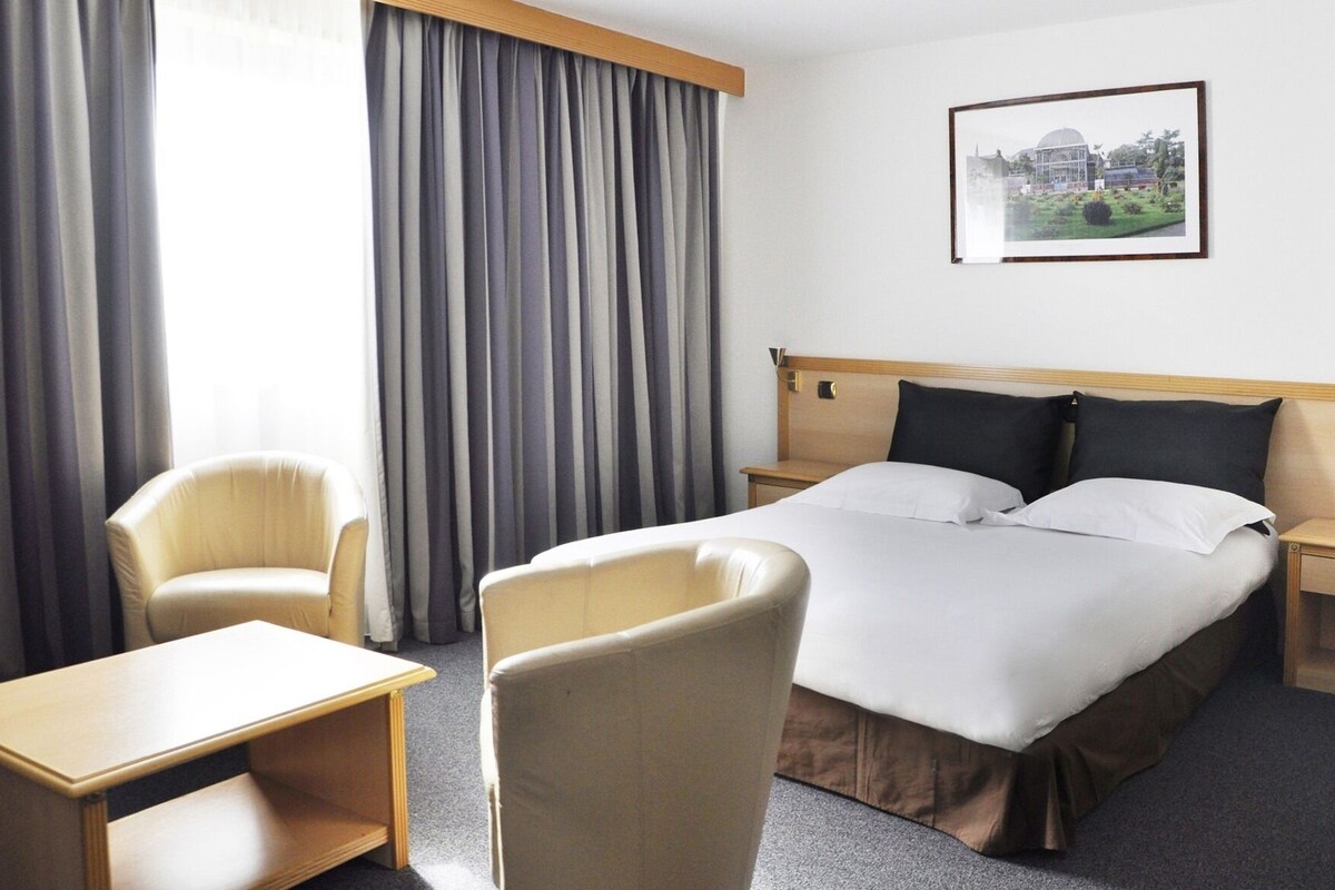 Une chambre d’hôtel avec un lit, deux chaises, un bureau, une table de chevet et un tableau encadré au mur.