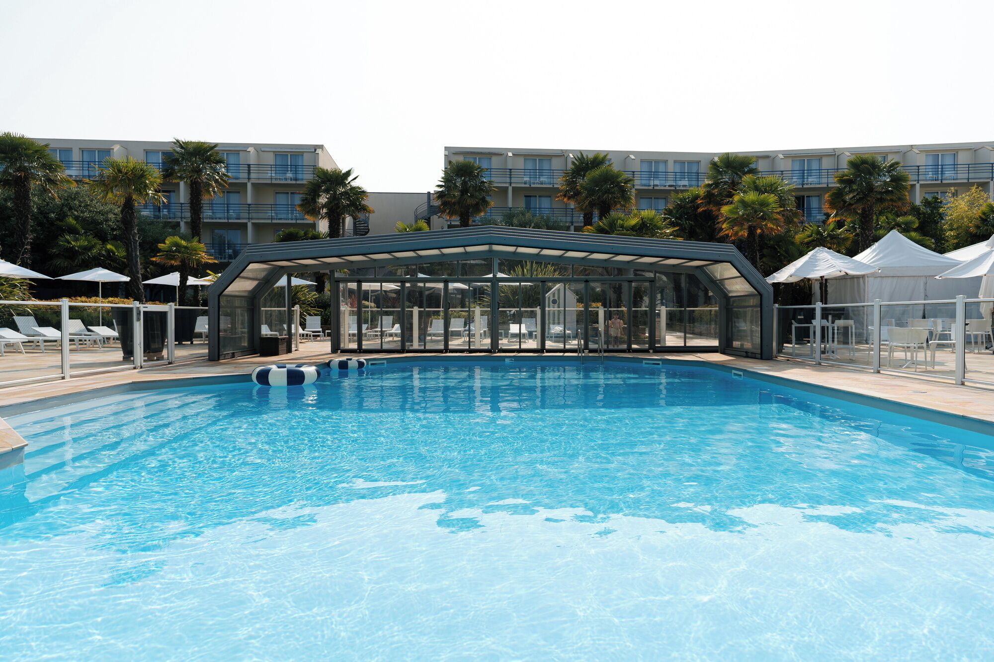A swimming pool with a glass enclosure, surrounded by lounge chairs and umbrellas, with a building in the background.