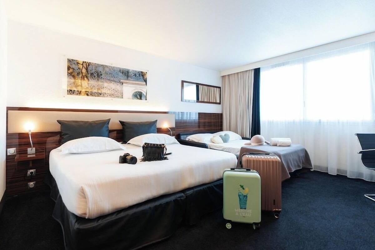 A hotel room with two beds, a large window, a painting on the wall, and a suitcase on the floor.
