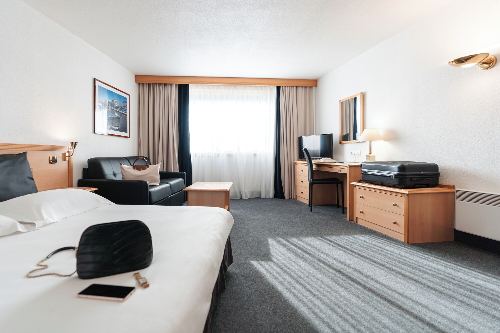 A hotel room with a bed, a desk, a chair, a TV, a lamp, and a suitcase.
