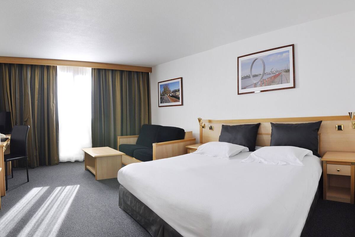 A hotel room with a large bed, a desk, a chair, a TV, and two framed pictures on the wall.