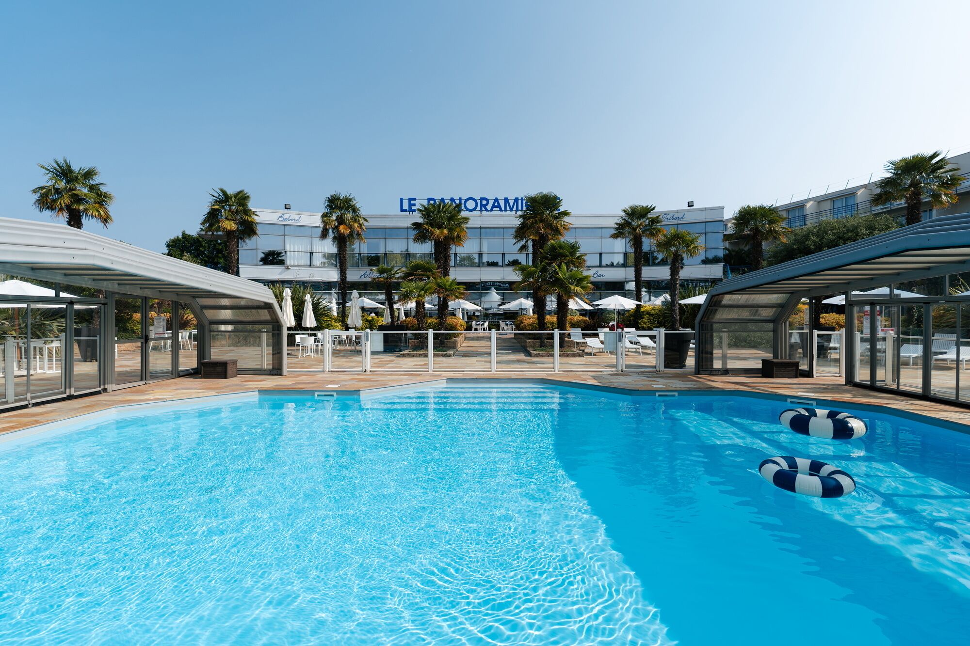 Ein Swimmingpool mit Liegestühlen und Sonnenschirmen, umgeben von Palmen und einem Hotel im Hintergrund.