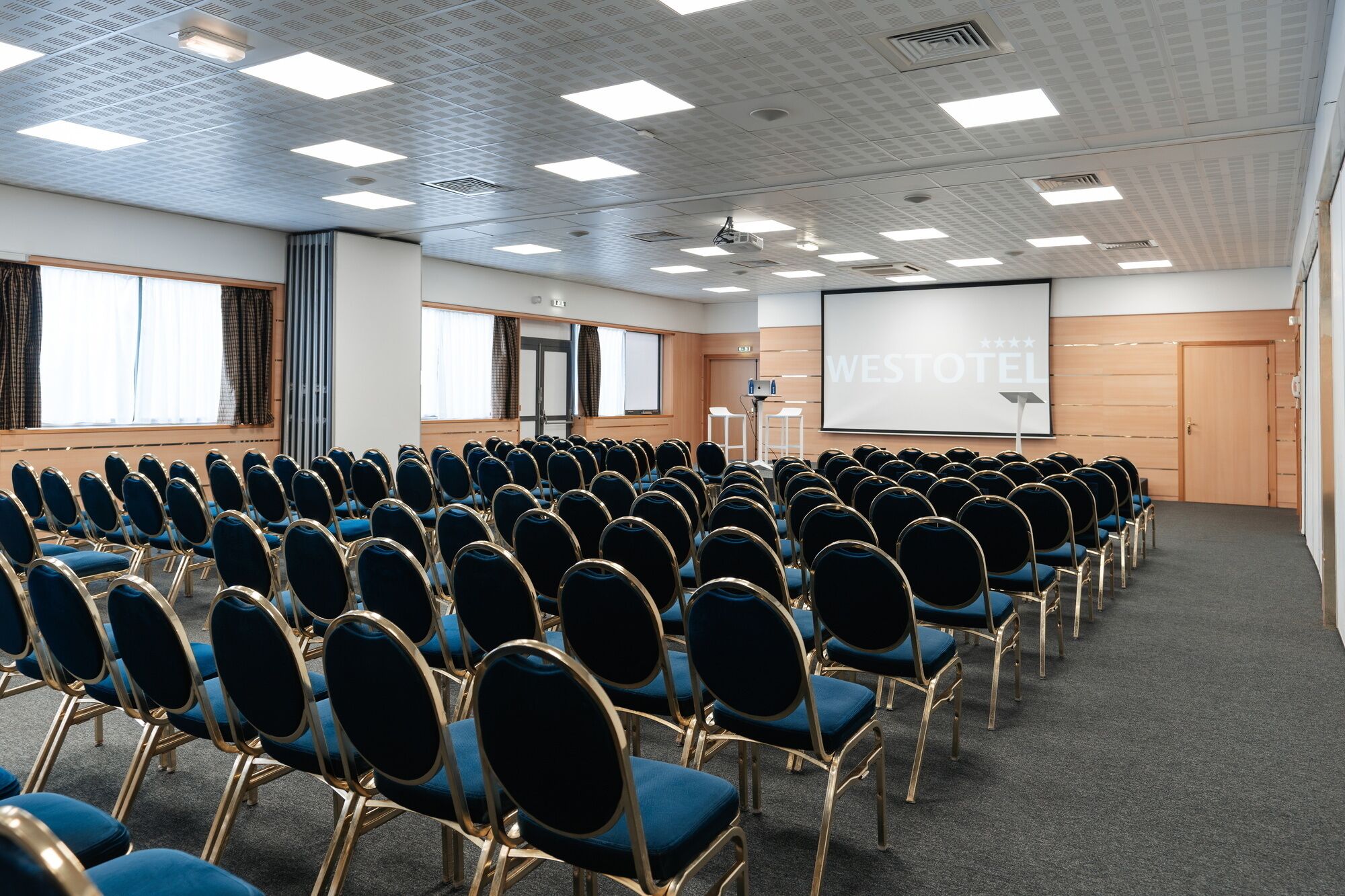 A conference room with rows of blue chairs, a projector screen, and a large screen displaying "WESTOTEL".