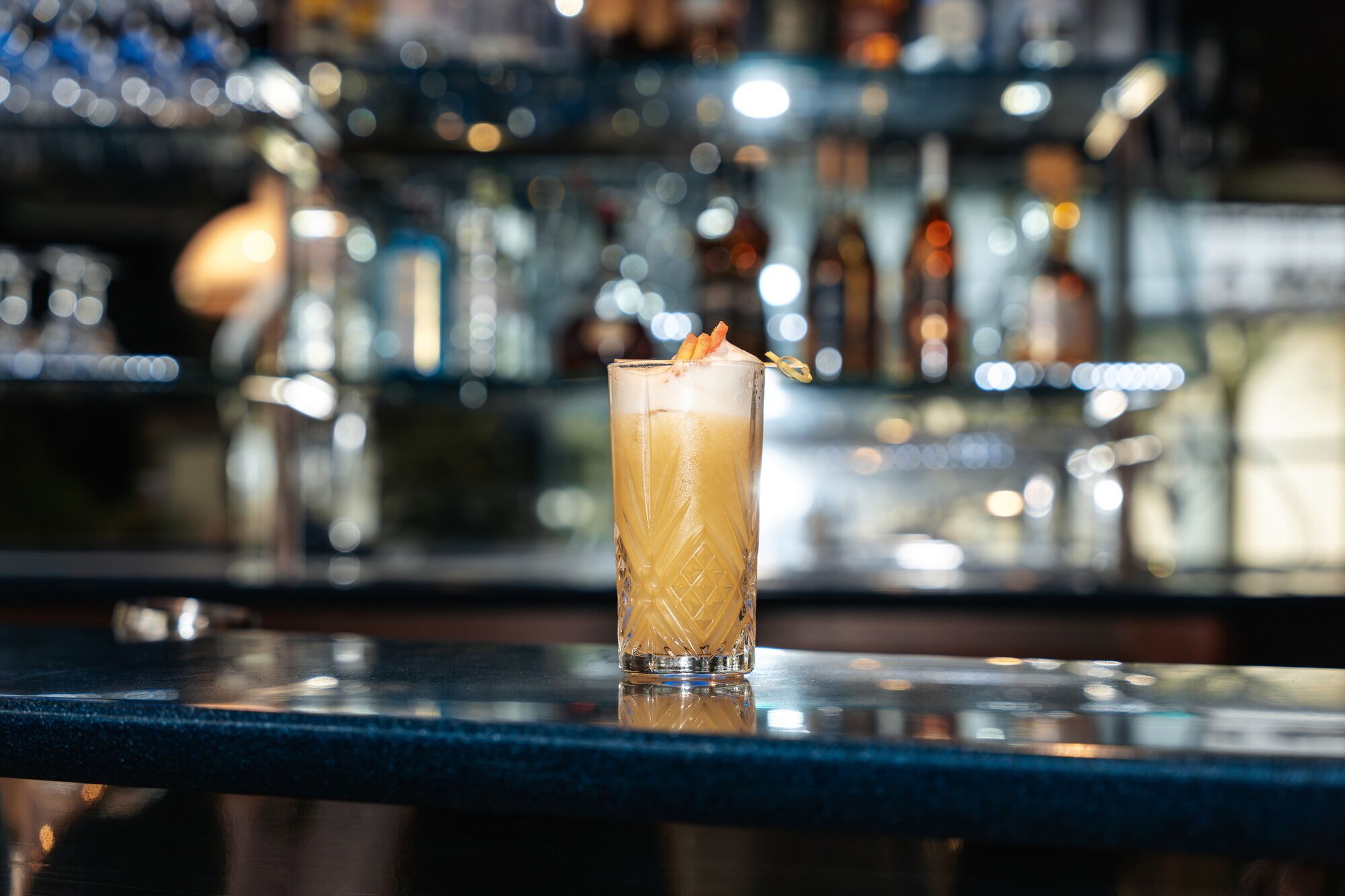 A bar setting with a tall glass of a layered drink, ice, and a garnish, with a blurred background of shelves with bottles.