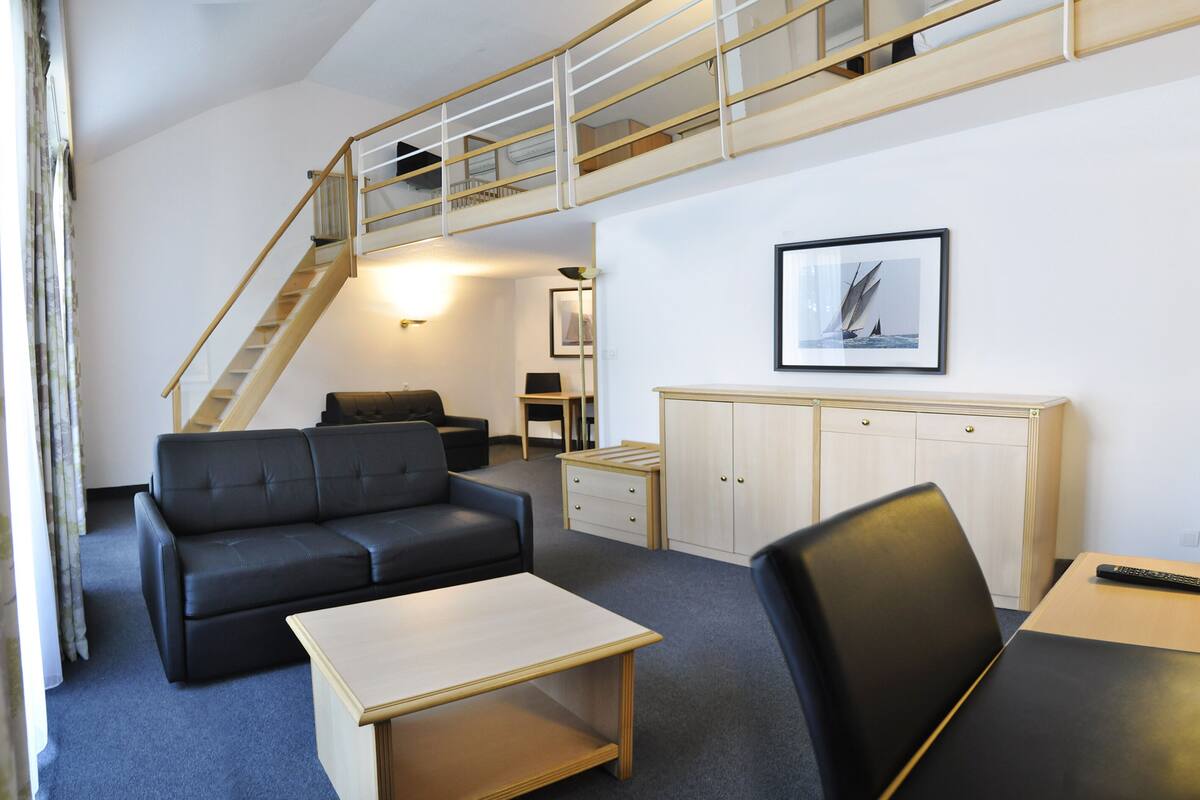 A compact living space with a sofa, a small table, a desk, a chair, a wardrobe, and a staircase leading to an upper level.