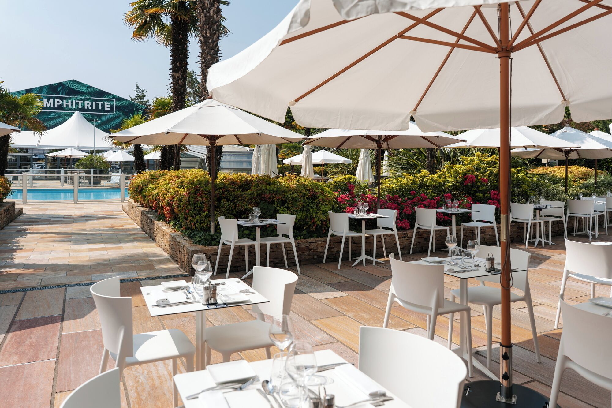 An outdoor dining area with white tables and chairs, surrounded by lush greenery and a pool in the background.