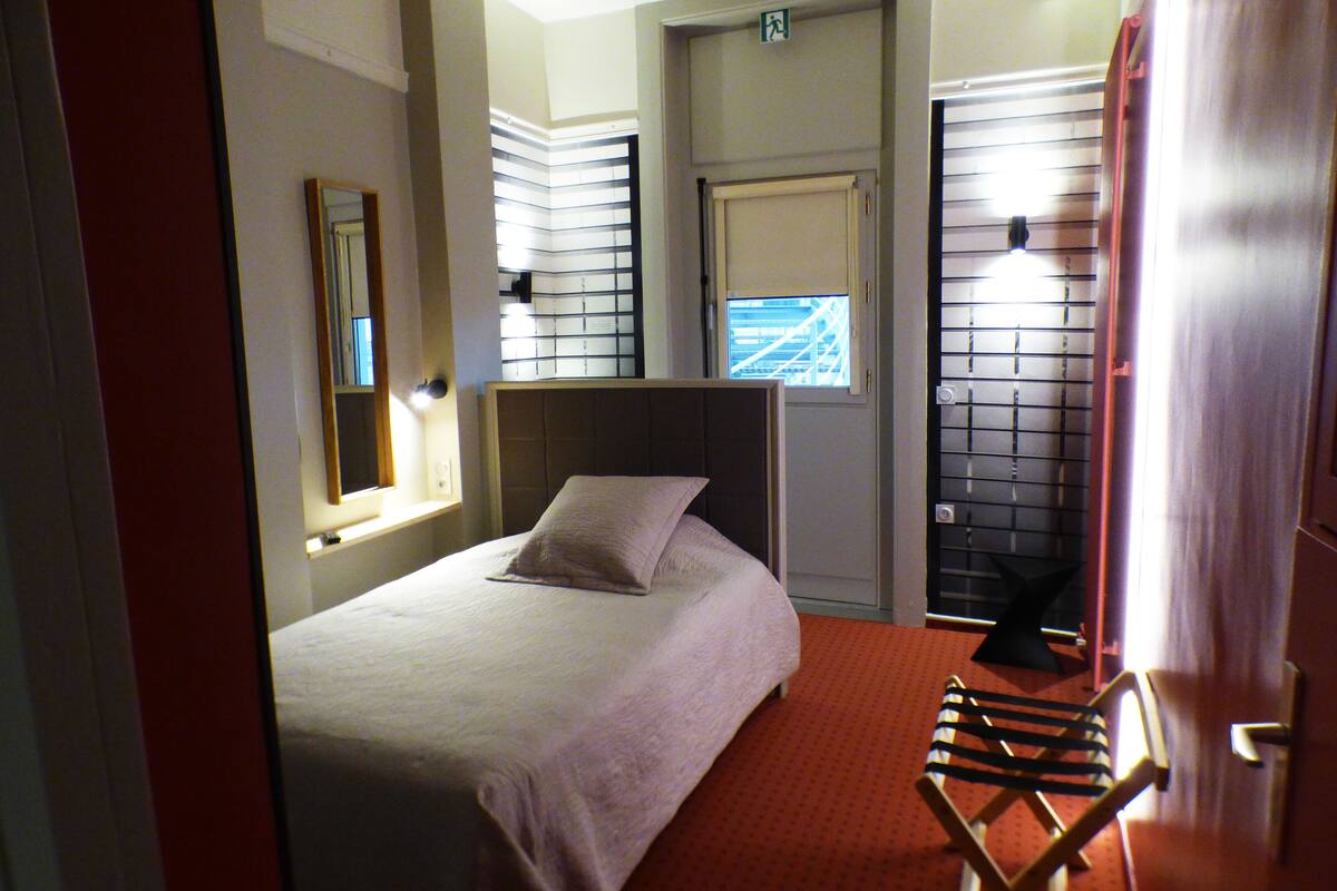 Habitación de hotel con cama, ventana con persianas, espejo, puerta y una silla.