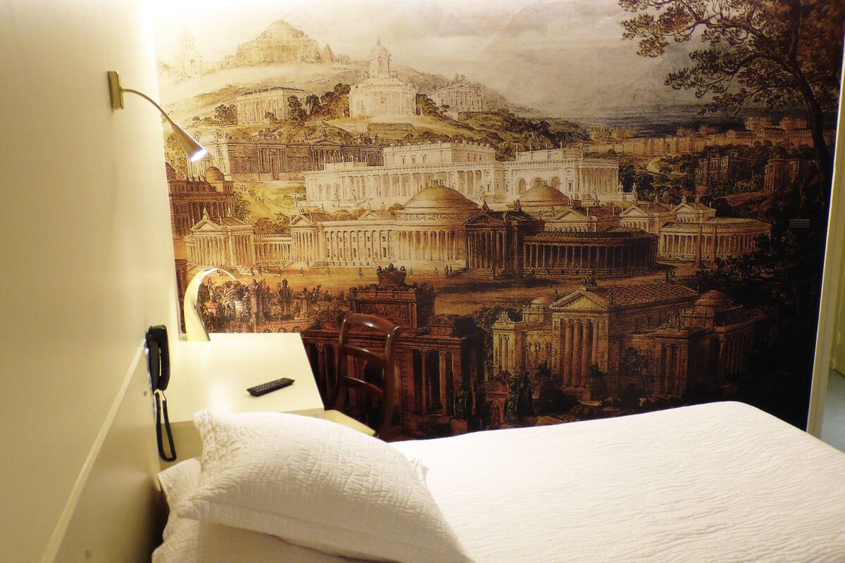 Habitación de hotel con cama, lámpara y un mural en la pared que muestra arquitectura antigua.