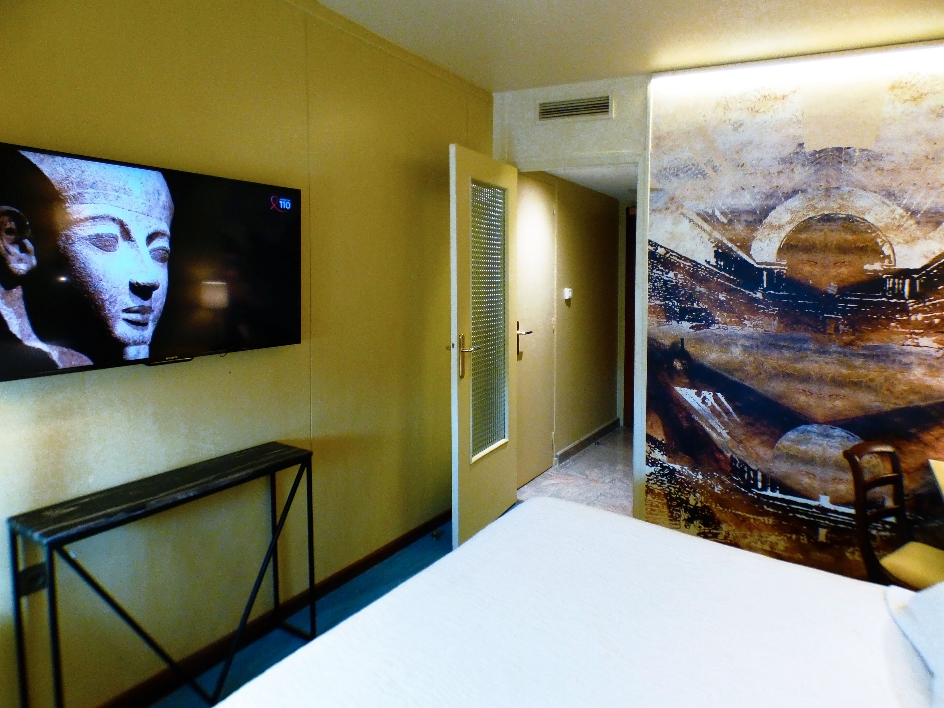 Habitación de hotel con una televisión montada en la pared, una puerta que da a otra habitación y un cuadro grande en la pared.