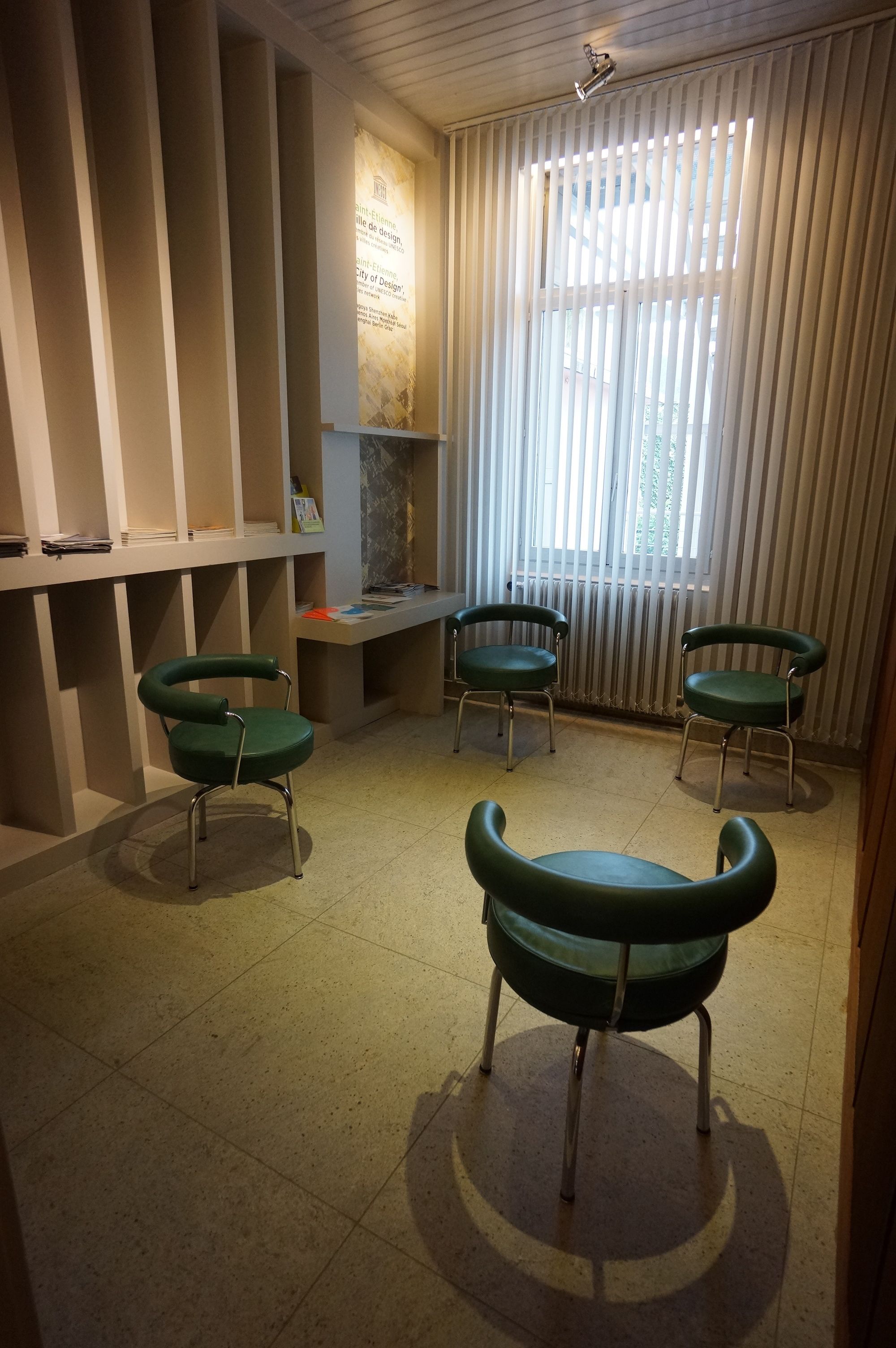 A room with a tiled floor, a desk with a computer, two green chairs, and a window with blinds.