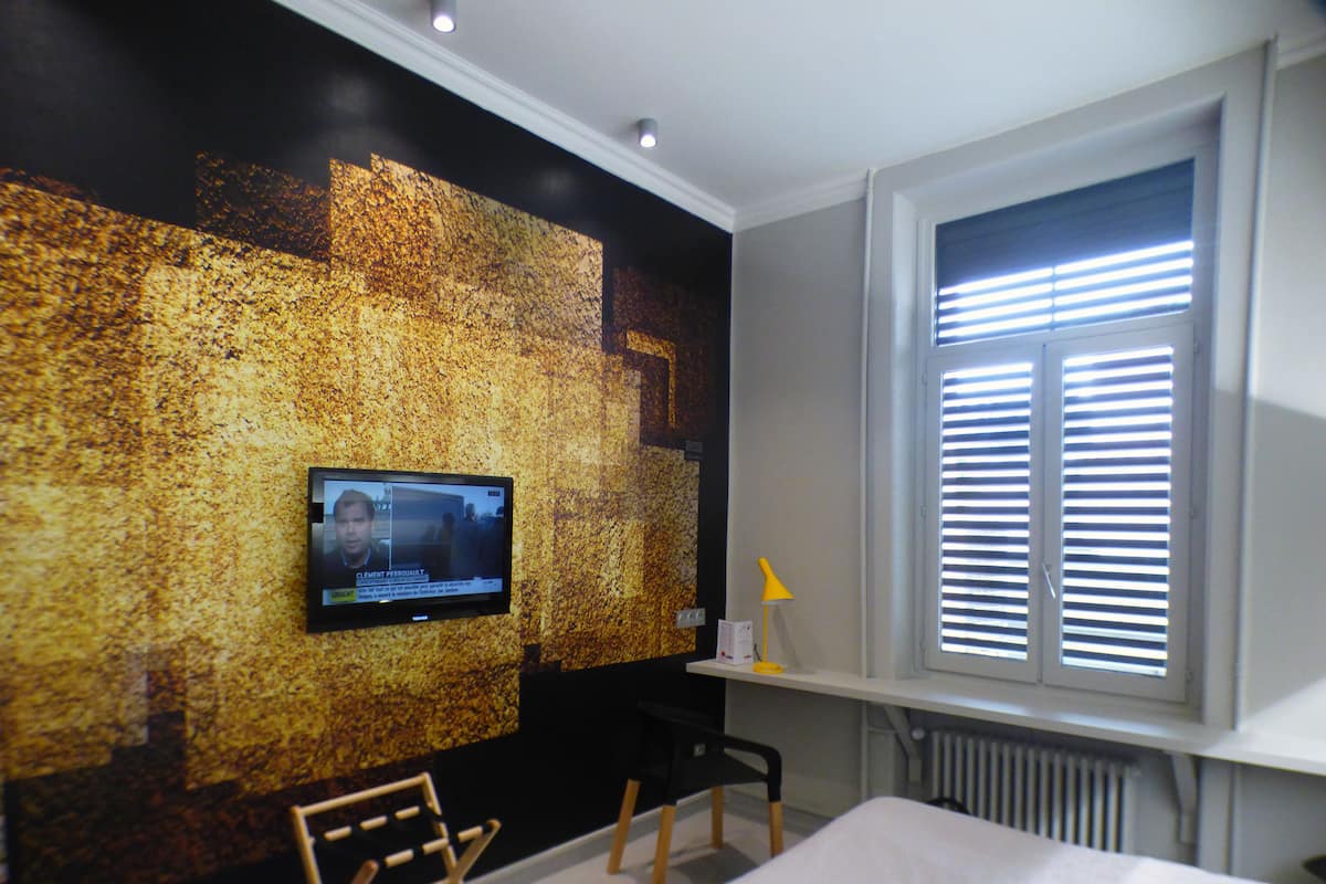 Una habitación de hotel moderna con una televisión montada en una pared texturizada, una ventana con persianas, un escritorio con lámpara y una cama.