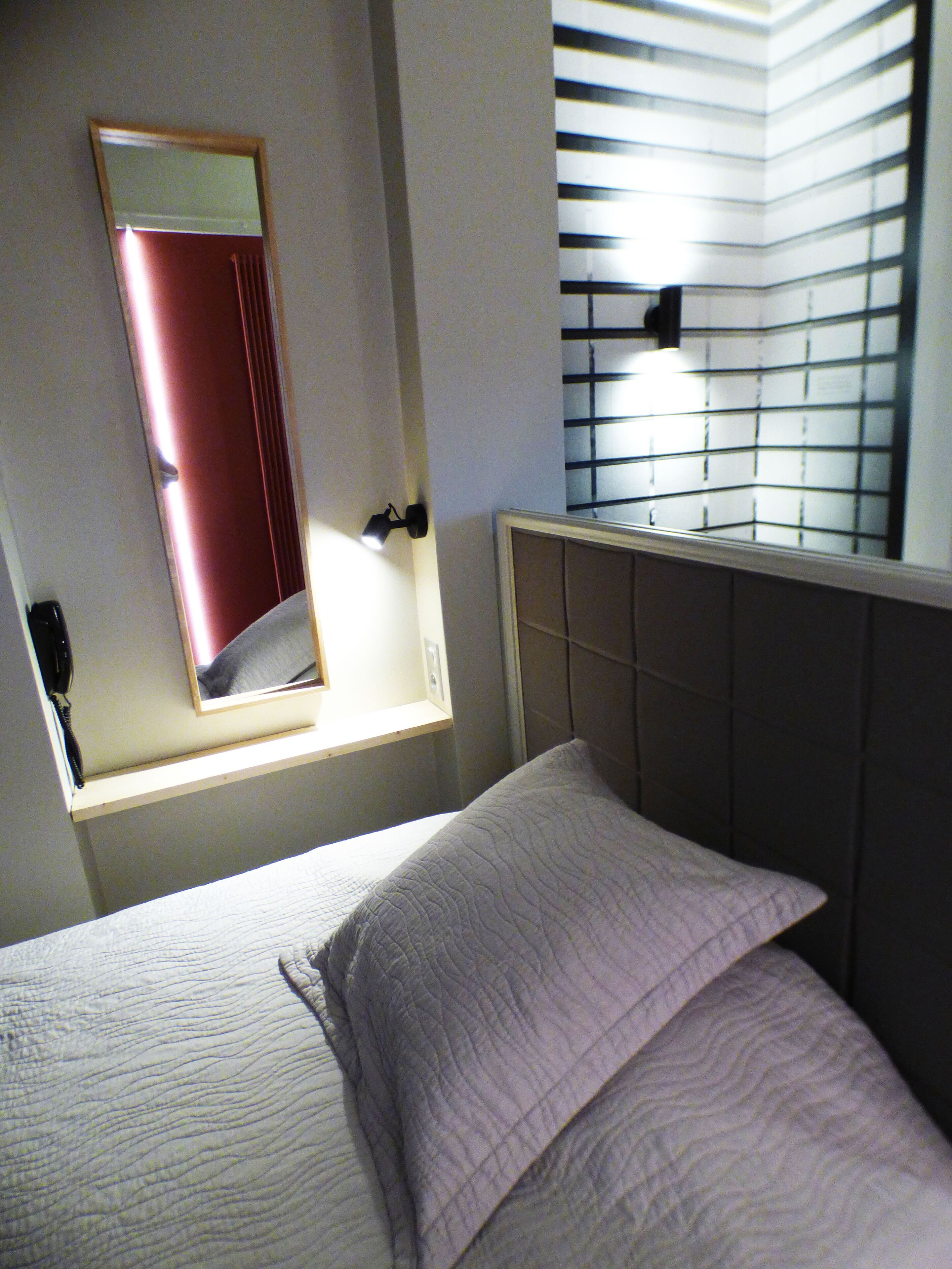 Un dormitorio con cama, lámpara de noche, ventana con persianas y espejo en la pared.