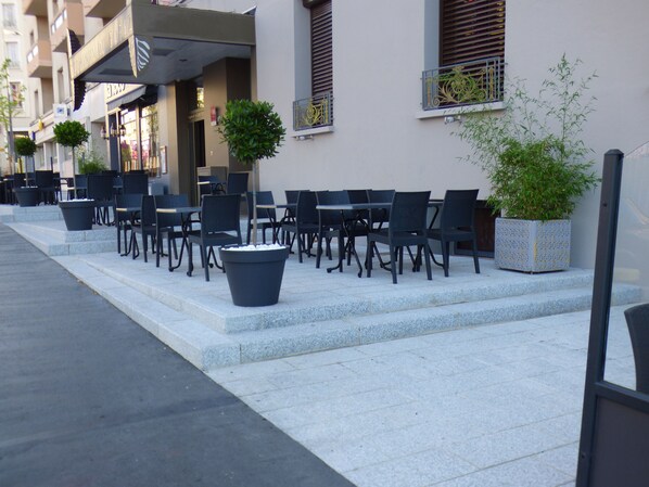 An outdoor seating area with tables and chairs, potted plants, and a building with a sign.
