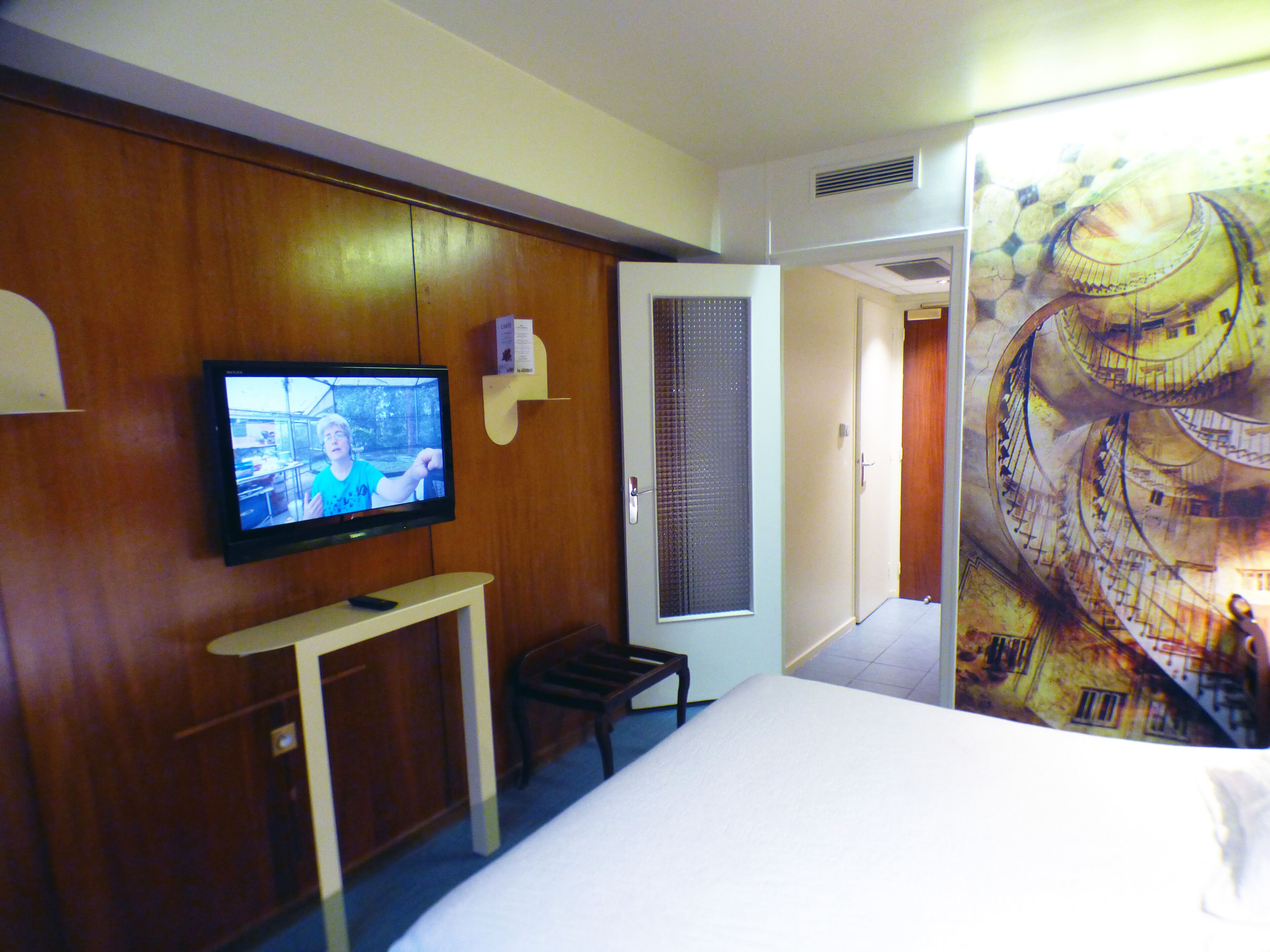 Habitación de hotel con una televisión sobre un mueble de madera, una cama, una mesita y una puerta que da a otra habitación.