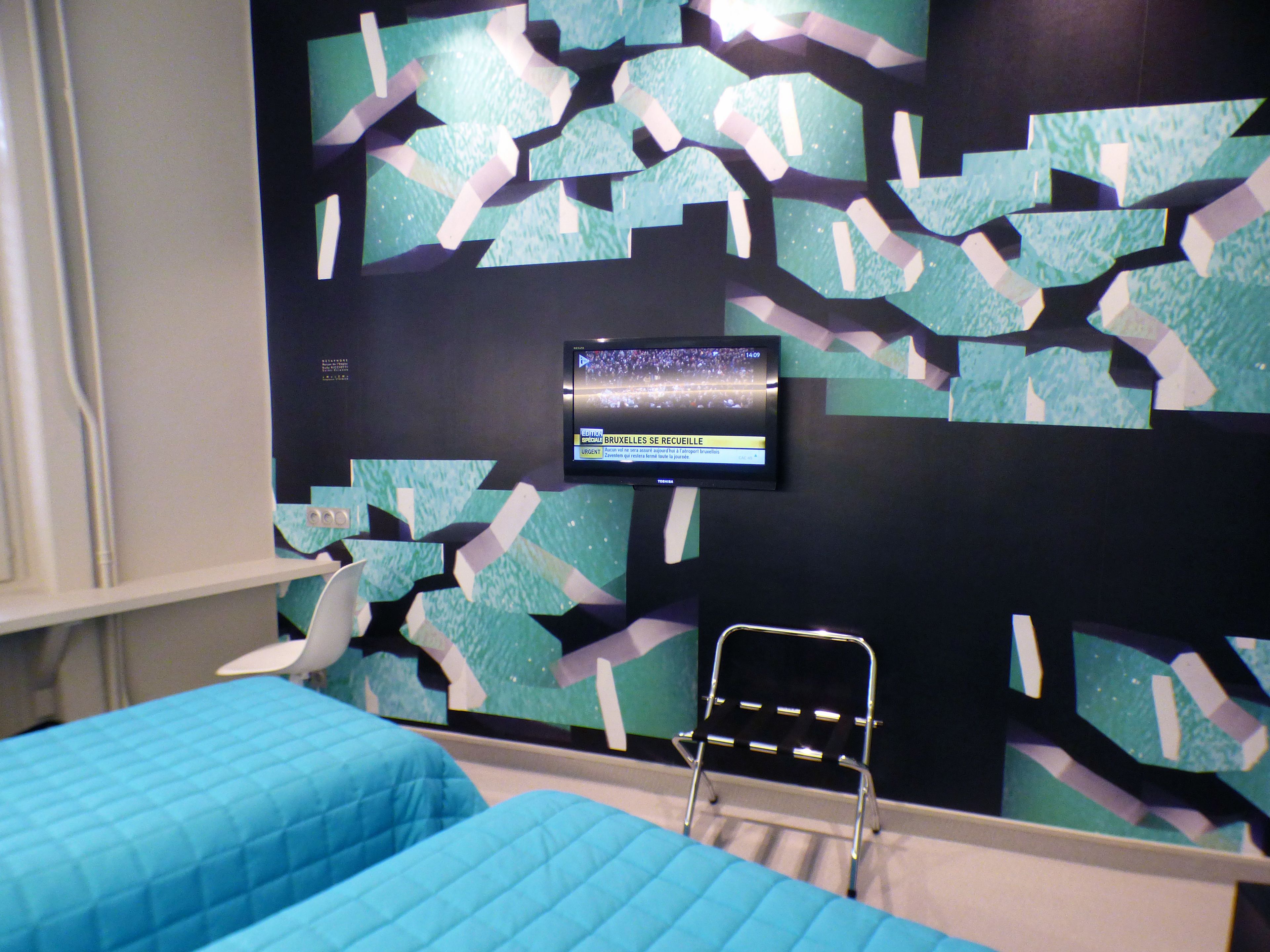 Een hotelkamer met een bed, een stoel en een televisie aan de muur, waarop een nieuwskanaal wordt vertoond.
