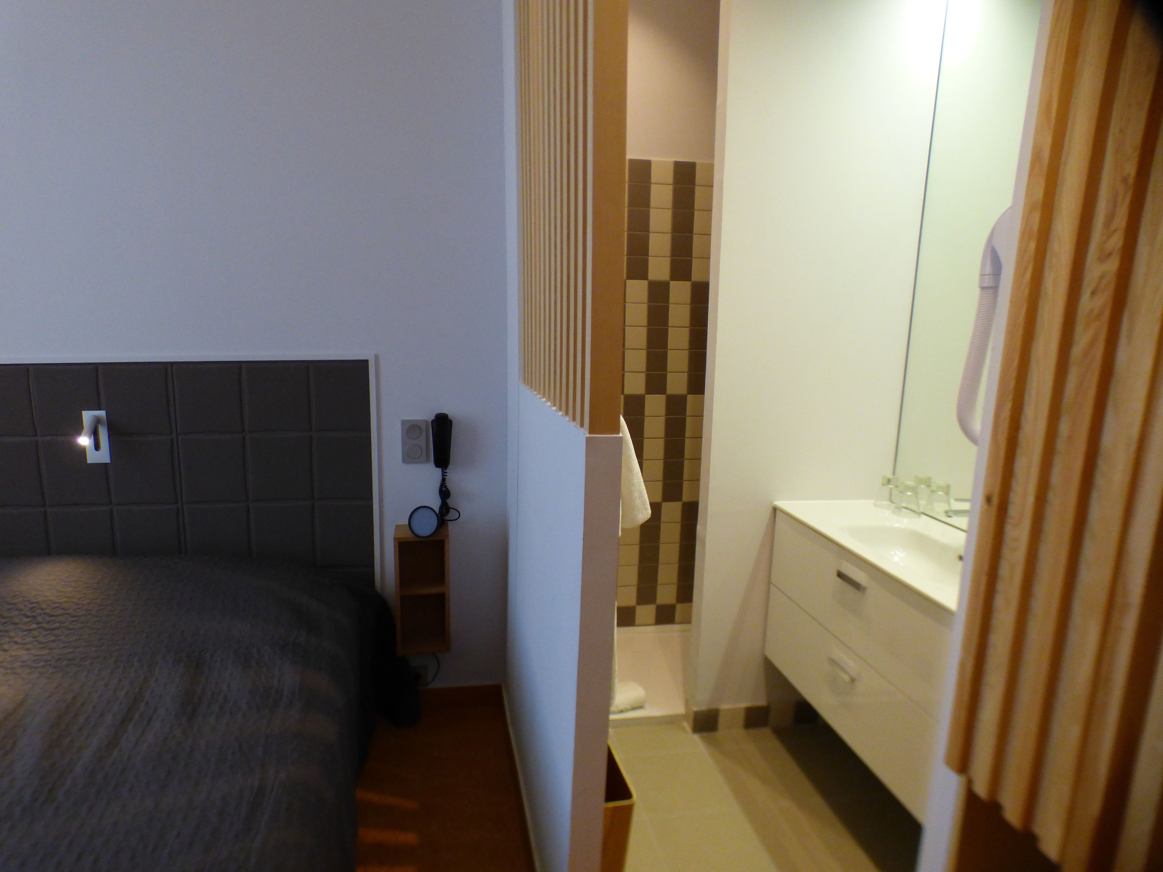 Une chambre d’hôtel comprenant un lit, un téléphone, une petite étagère en bois et une salle de bain avec un miroir, un lavabo et un porte-serviettes.