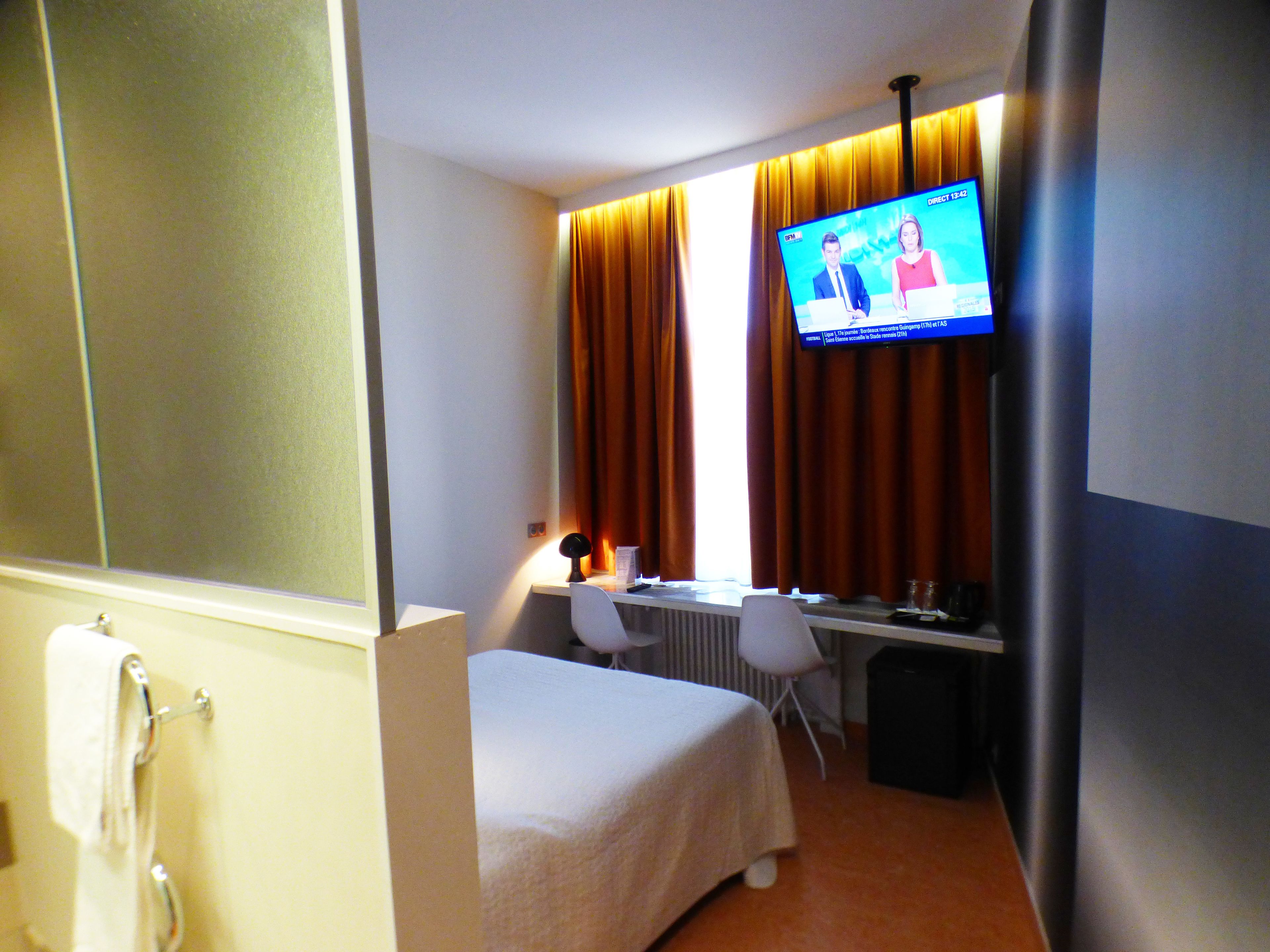 Ett hotellrum med en säng, ett skrivbord, en stol, en tv och ett fönster med gardiner.