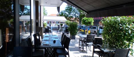 An outdoor cafe with tables and chairs, a covered seating area, and a view of the street.