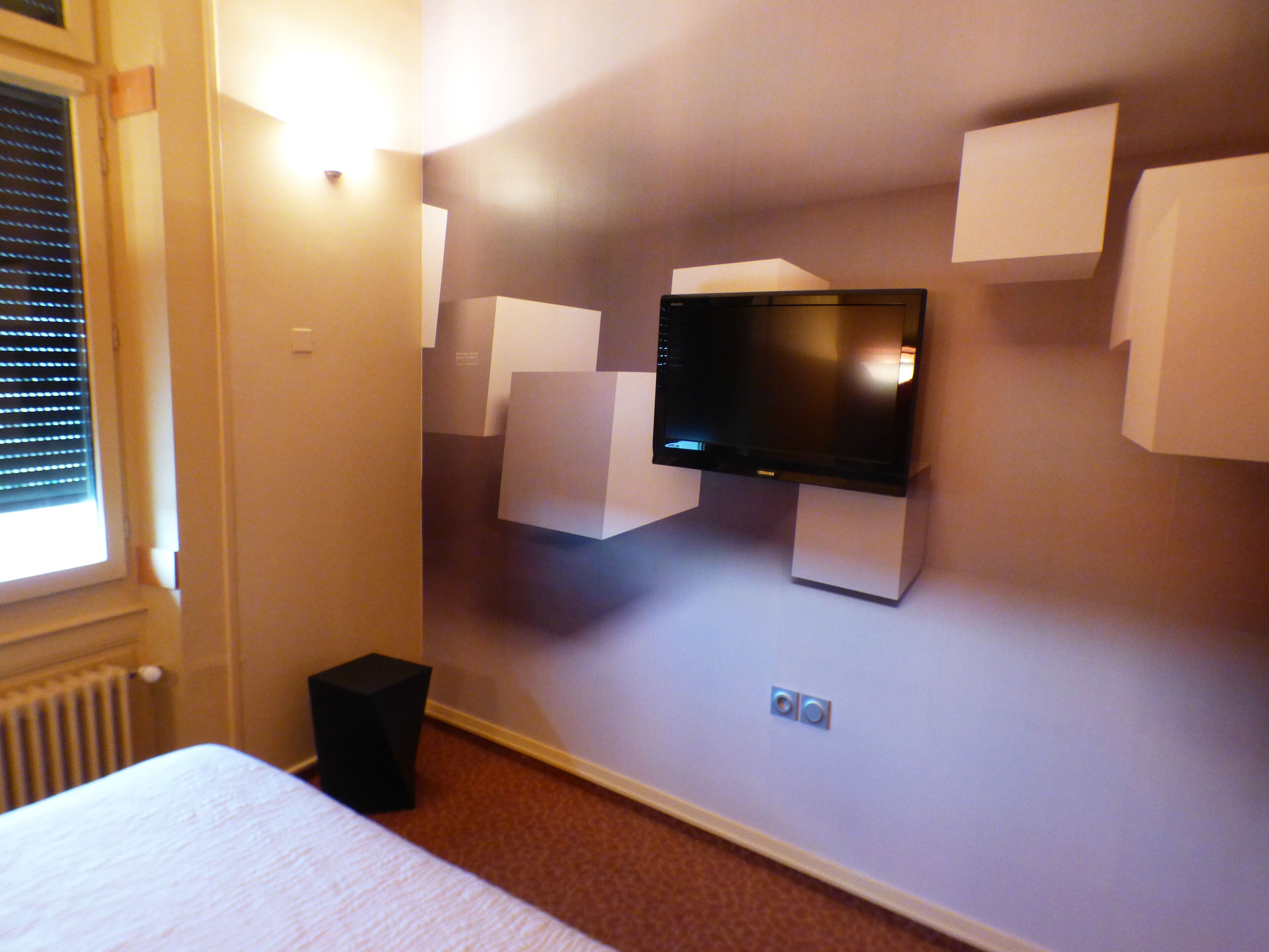 A hotel room with a flat-screen TV mounted on the wall, a bed, and a small black speaker.