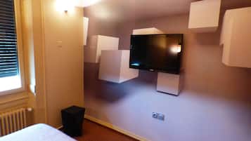 A hotel room with a flat-screen TV mounted on the wall, a bed, and a small black speaker.