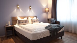 Een keurig opgemaakt bed met witte lakens en een grijze deken, een houten nachtkastje met een witte kaars, en een stoel met een gestreept kussen.