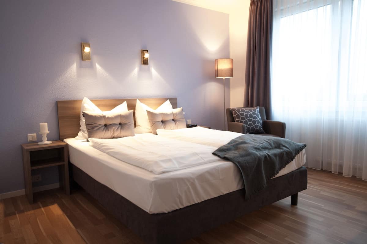 A neatly made bed with white linens and a gray throw blanket, a wooden nightstand with a white candle, and a chair with a patterned cushion.