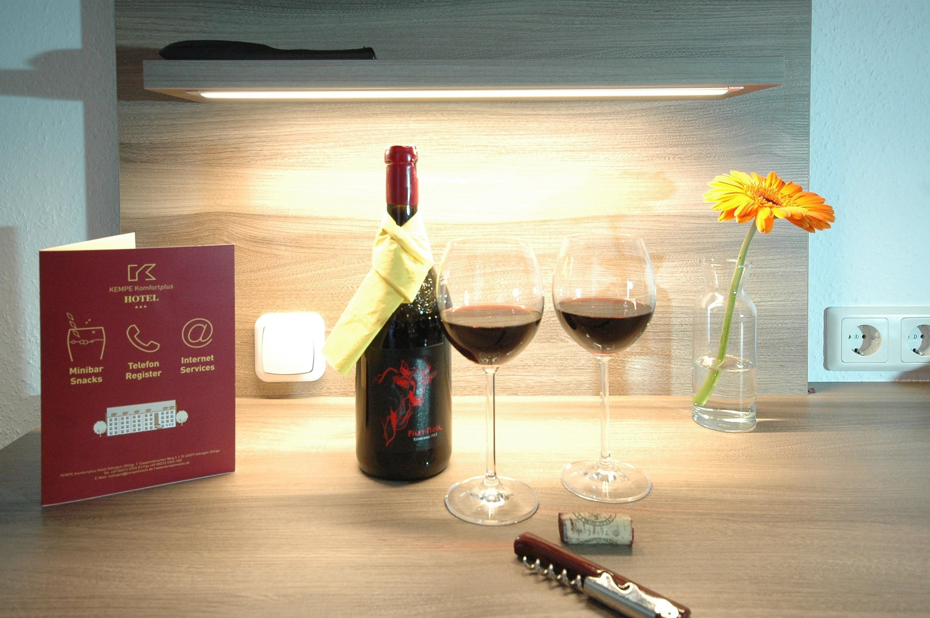 Un mini-bar d’hôtel contenant une bouteille de vin, deux verres à vin, une fleur dans un vase et un tire-bouchon, le tout posé sur une surface en bois.