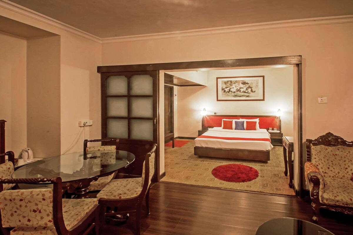 Ein Hotelzimmer mit einem Glastisch, Holzsesseln, einem Bett mit roter Bettwäsche und einem roten Teppich.
