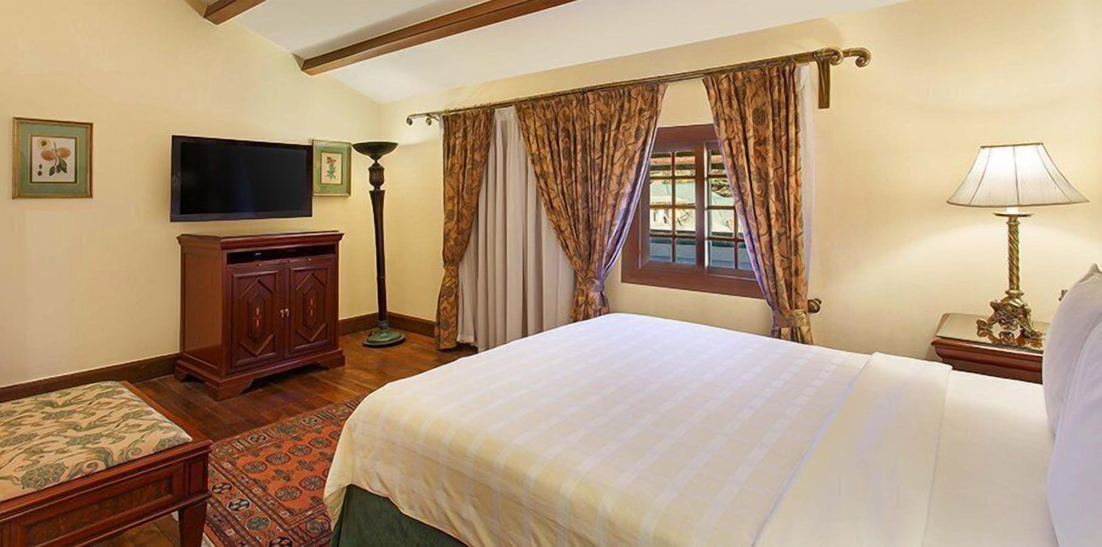 A hotel room with a large bed, a television, a desk with a lamp, and a window with curtains.
