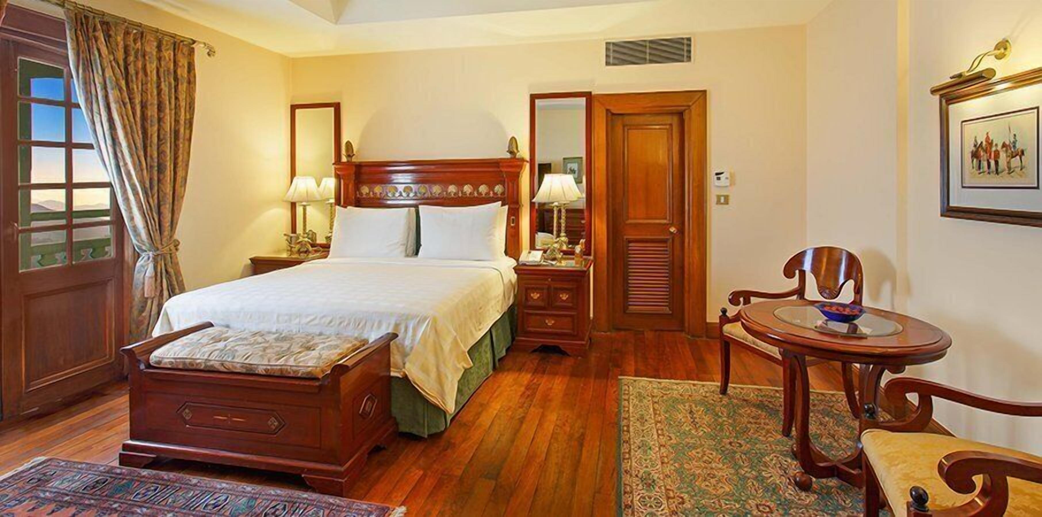 A hotel room with a wooden bed, a small round table, a chair, a nightstand, a lamp, a mirror, and a door with a view.