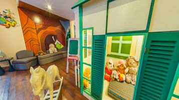 Una sala de juegos infantil con piso de madera, mobiliario colorido y una casita de juegos con contraventanas verdes.