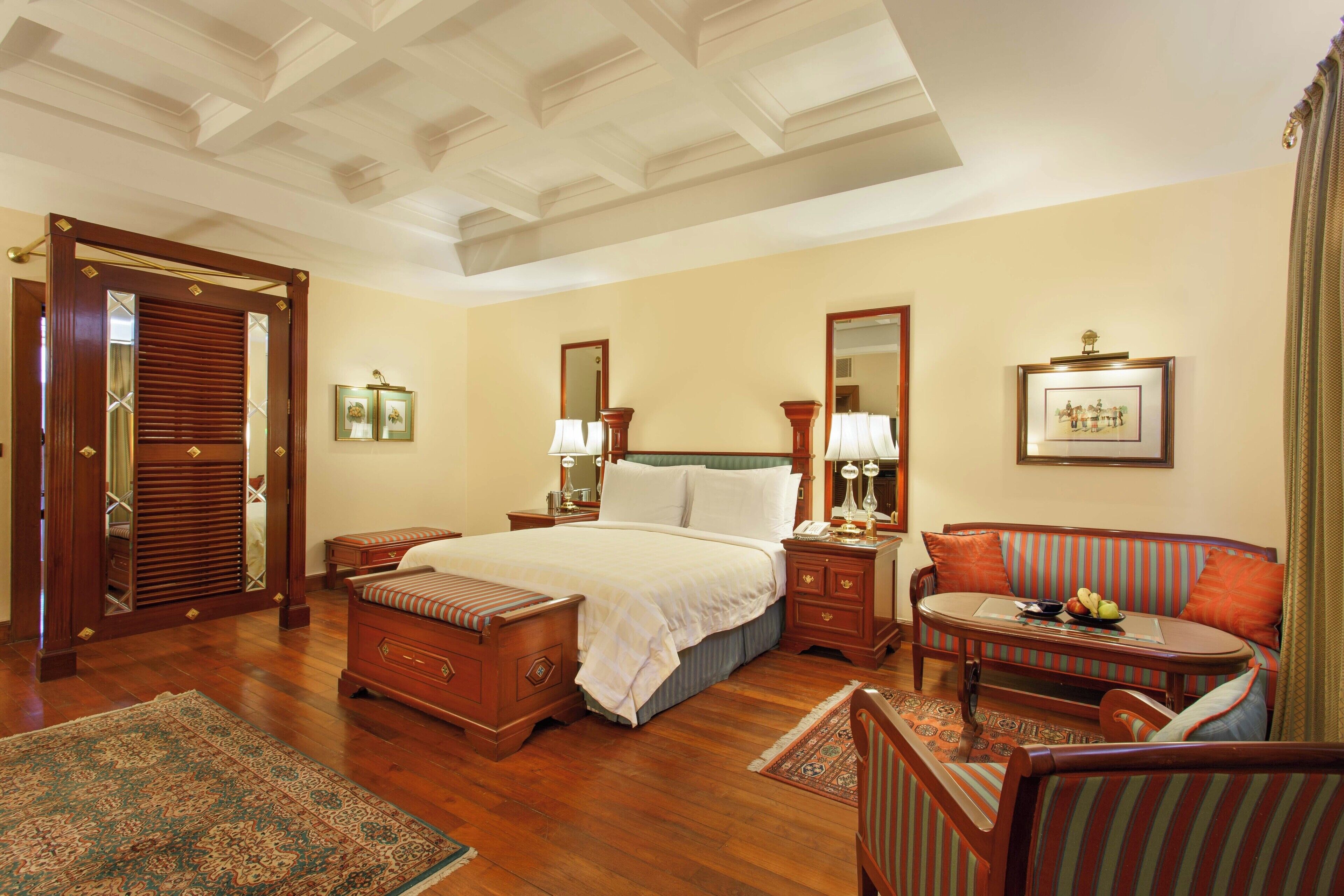 A spacious bedroom with a large bed, a sitting area with a sofa and armchairs, a wooden floor, and a ceiling with a decorative design.