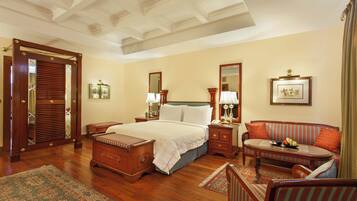 A spacious bedroom with a large bed, a sitting area with a sofa and armchairs, a wooden floor, and a ceiling with a decorative design.