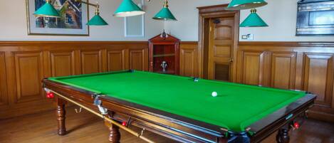 A pool table with a green felt surface, wooden surroundings, and a chandelier with green lamps.