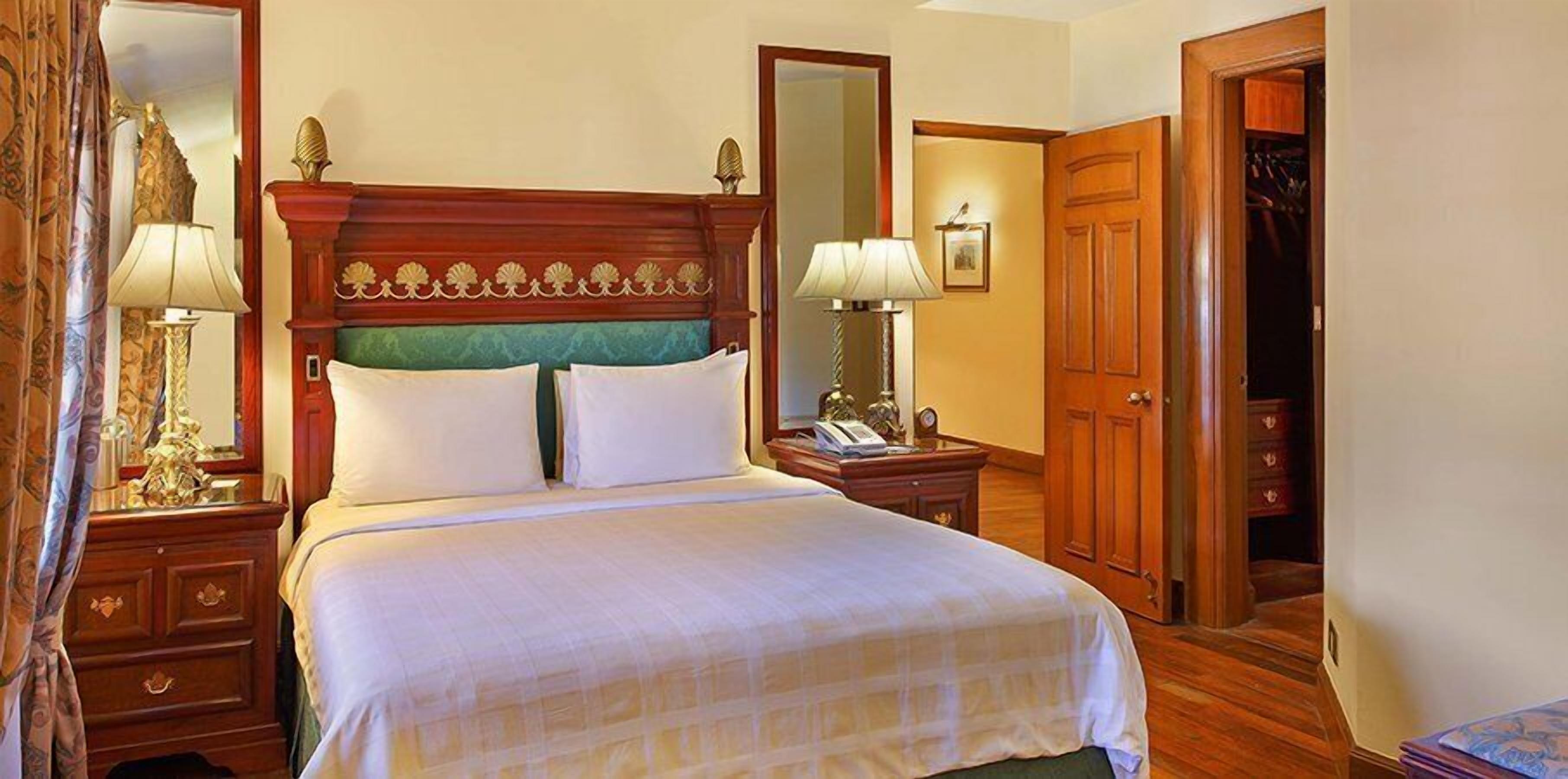 A hotel room with a large bed, two bedside tables, a nightstand, a wooden wardrobe, and a mirror.