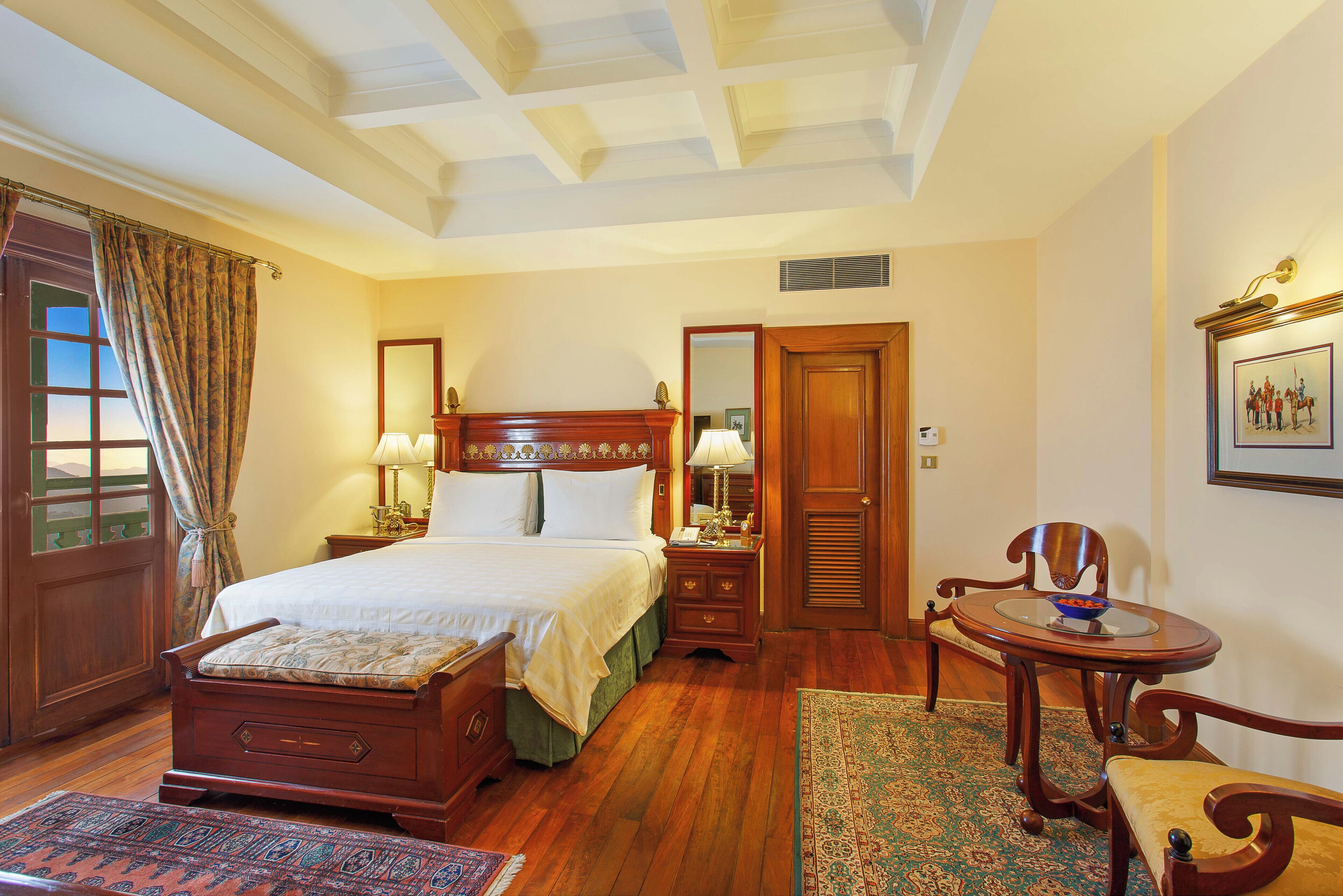 A traditional bedroom with a wooden bed, a small round table with a chair, a mirror, and a framed picture on the wall.