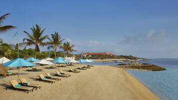 A beach with sun loungers and umbrellas, a hotel in the background, and boats on the water.