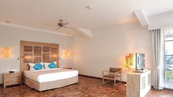 A bedroom with a large bed, wooden headboard, bedside lamps, a ceiling fan, a TV on a stand, and a view of the outdoors through a window.