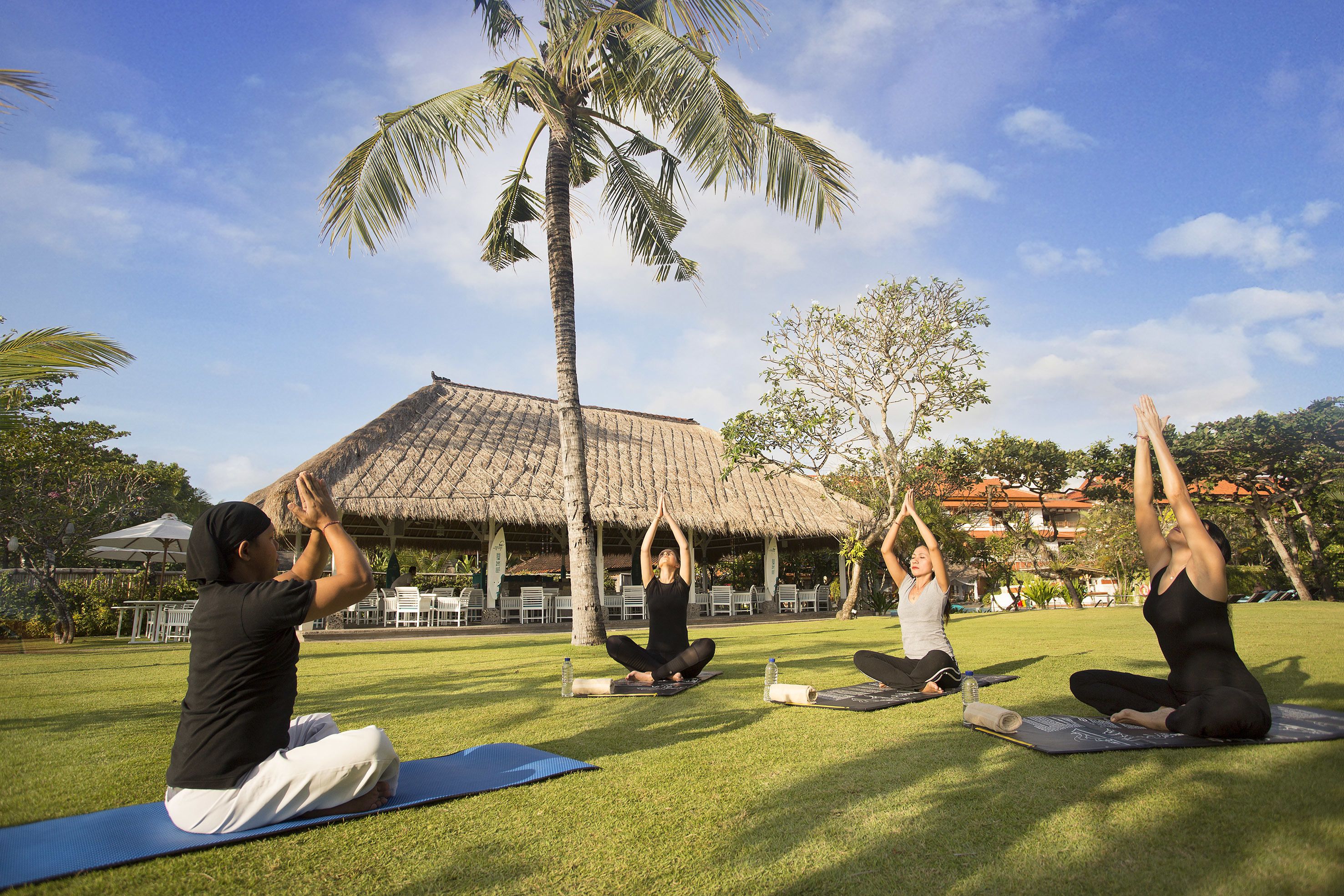Um grupo de pessoas praticando yoga em tapetes, em ambiente externo, com uma estrutura de telhado de palha ao fundo.
