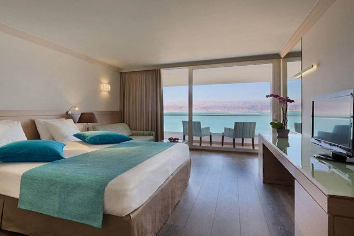 A hotel room with a bed, a TV, a balcony with a view of the sea, and a desk with a chair.