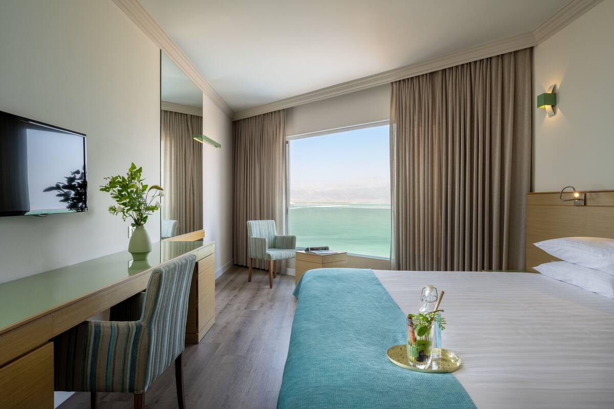 A hotel room with a bed, a desk, a chair, a television, and a large window with a view of the sea.