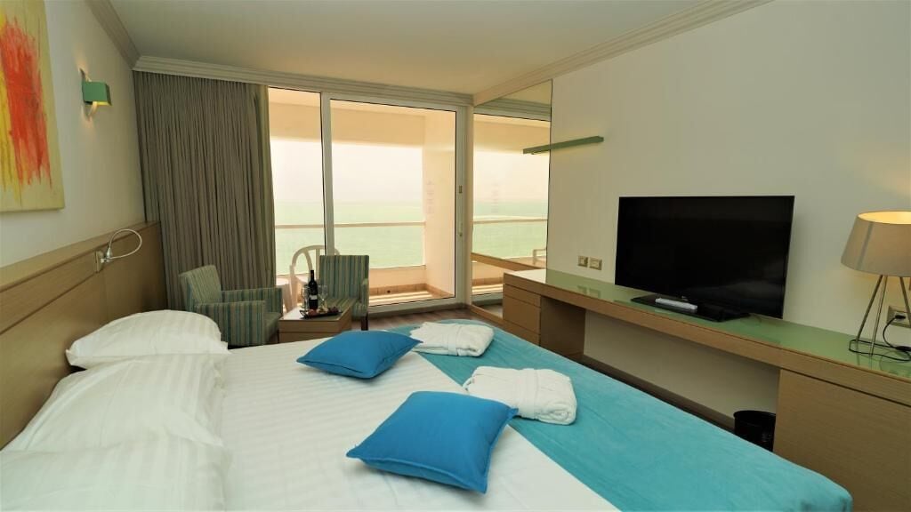 Quarto de hotel com cama, televisão, escrivaninha e vista para o mar.