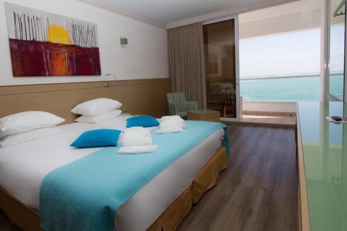 A hotel room with a bed, a view of the sea, a painting on the wall, and a balcony with a chair.