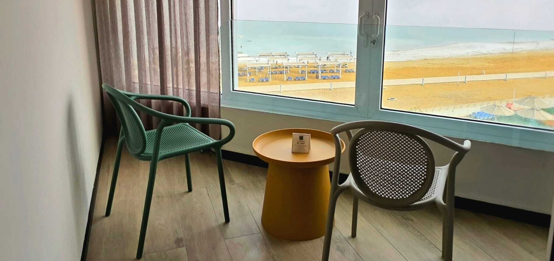 Una habitación con dos sillas, una mesa pequeña y una ventana que ofrece vista a una playa y coches estacionados.