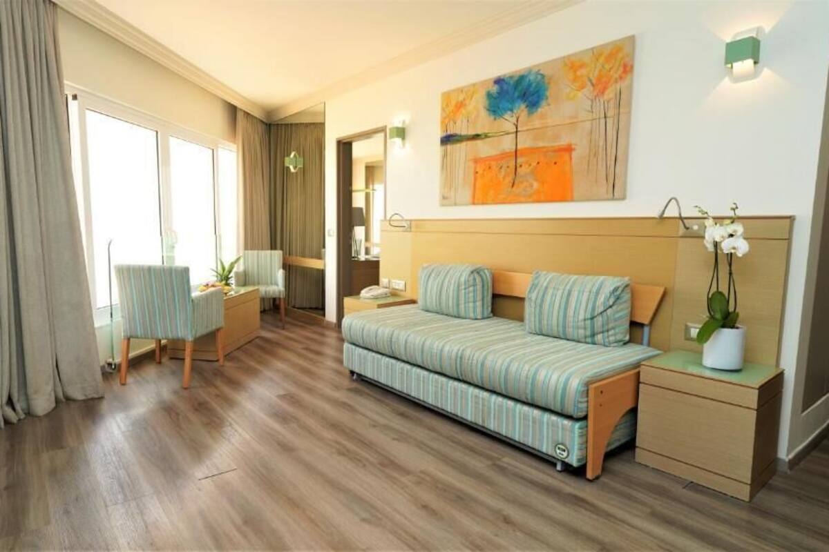 A hotel room with a sofa bed, a small table, a chair, a desk, and a painting on the wall.