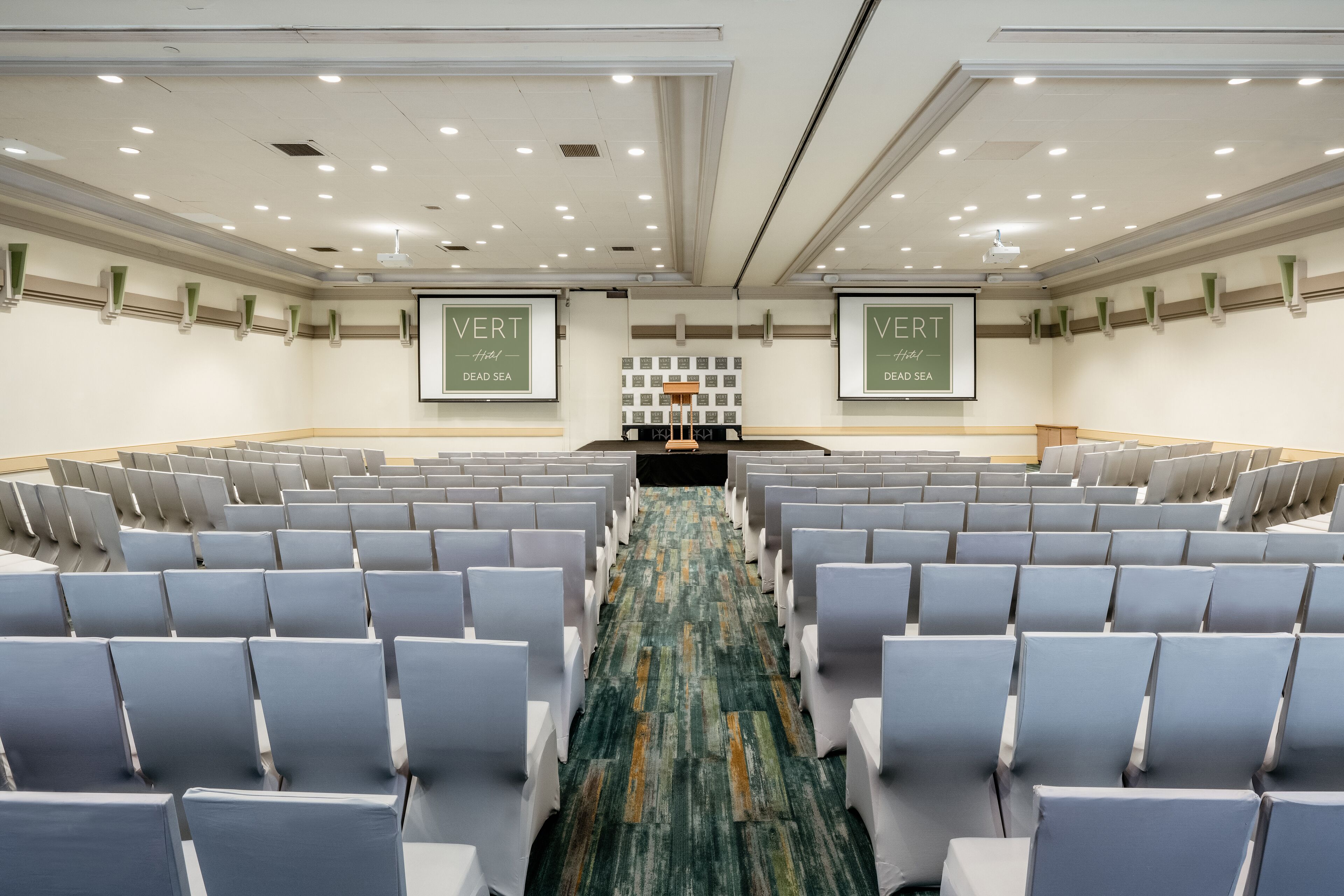 Un salón de conferencias con filas de sillas, un escenario y dos pancartas con la leyenda 'VERT' y 'DEAD SEA'.
